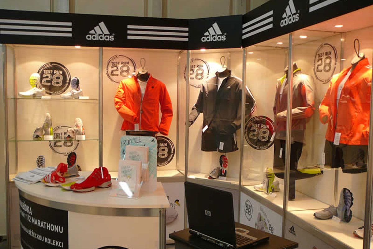 Výstavní expozice sportovních produktů značky Adidas Vitríny a stolek na výstavní expozici s produkty Adidas, límce stánku vylepené logy Adidas